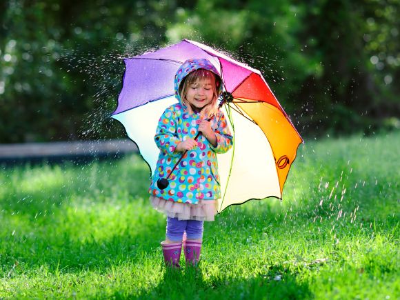 Young girl in a rain coat and wellies under a rainbow umbrella in the rain