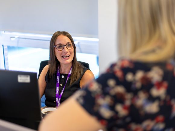 Smiling female member of Platform Housing Group staff sat at her desk in front of a computer talking to a out of focus female member of staff stood at the front of the image