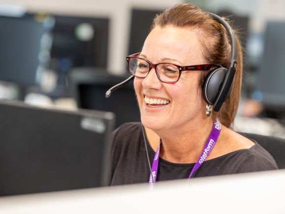 Colleague wearing a headset and Platform lanyard smiles while working at a desk in an open-plan office.