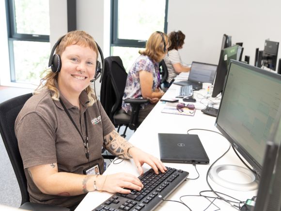 Female staff member sat at a desk working on a computer looking up and smiling at the camera.