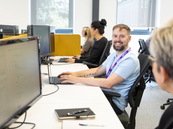 Open-plan office scene with colleagues working at desks; one person in the foreground smiles towards the camera while using a laptop.
