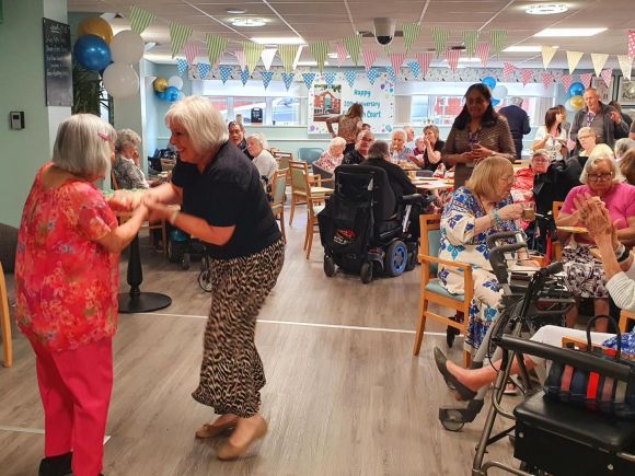 Older residents gather in a decorated communal room for a celebration. Two women dance together in the centre while others sit nearby watching, chatting, and clapping, with some using wheelchairs or walking frames.