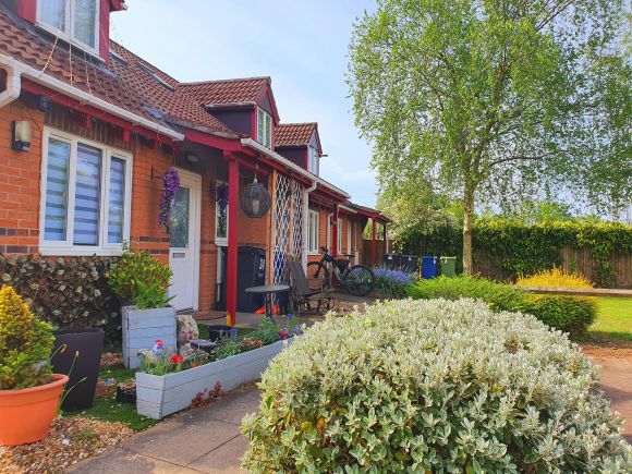 A row of single-storey red-brick bungalows with front gardens, a bench, and a paved pathway.