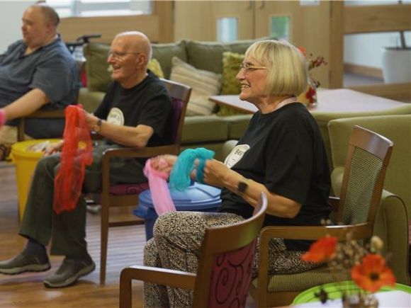 Three older adults seated indoors taking part in a group activity, holding colourful fabric scarves.