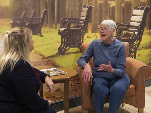 An older woman sitting in an armchair talking with a staff member in a lounge area, with a mural of a park scene behind them.