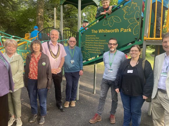 A group of adults stand in front of a new children’s playground structure in Whitworth Park, with several children playing on the equipment behind them. A large green sign on the play area shows the park name and funders, and the scene is set in a leafy park on a sunny day.
