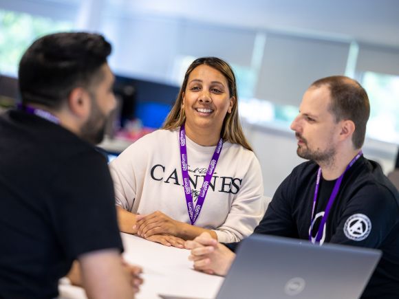 Three colleagues sitting at a desk in an office, having a discussion. One person is speaking while the other two listen, with a laptop open on the table. All are wearing purple Platform lanyards.