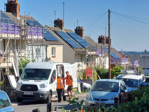 Construction workers beside a van on a residential street with scaffolding and solar panels being installed on rooftops.