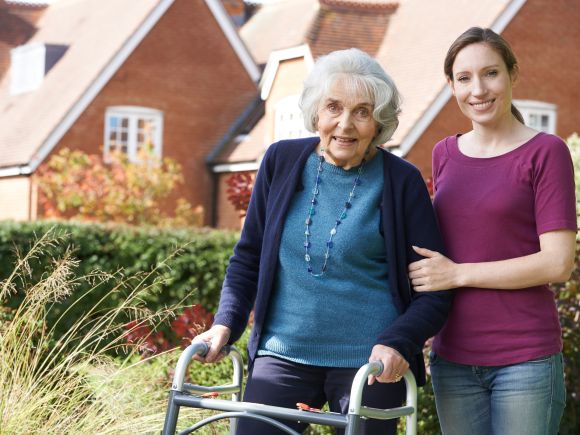 An older woman using a walking frame stands outside in a garden beside brick houses. A younger woman stands next to her with a supportive arm around her.