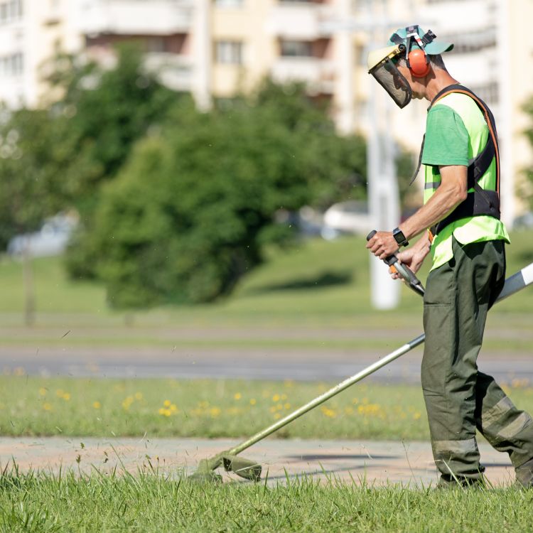 Male with high-vis clothing, face guard and safety equipment strimming a grassed area on an estate