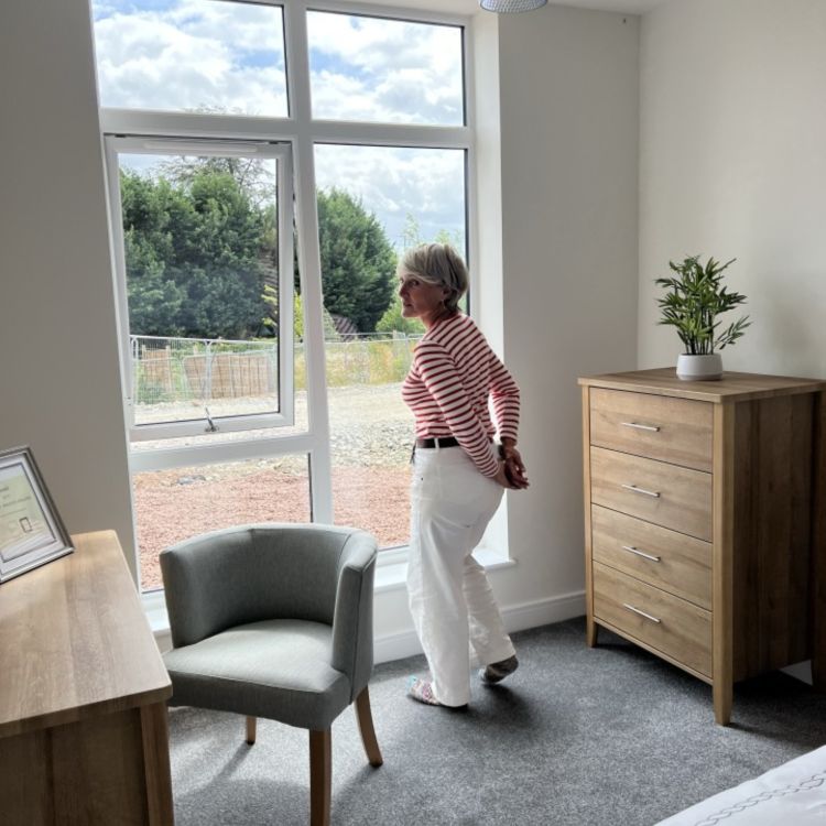 A woman stands in a bright, unfurnished bedroom, looking out through a large window. The room contains a chair, a chest of drawers, a desk and a bed, with light coming in from outside.