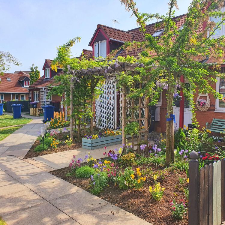 Wintern Court, Lincolnshire: A landscaped residential pathway lined with flowers, shrubs and small trees, leading past red-brick homes with pitched roofs.
