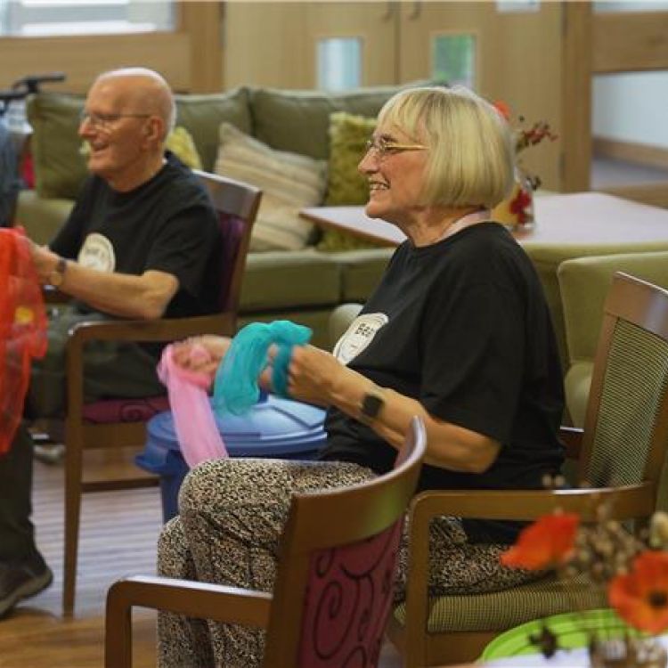 Three older adults seated indoors taking part in a group activity, holding colourful fabric scarves.