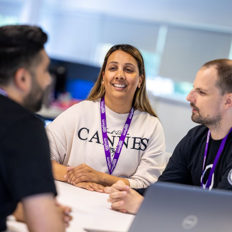 Three colleagues sitting at a desk in an office, having a discussion. One person is speaking while the other two listen, with a laptop open on the table. All are wearing purple Platform lanyards.