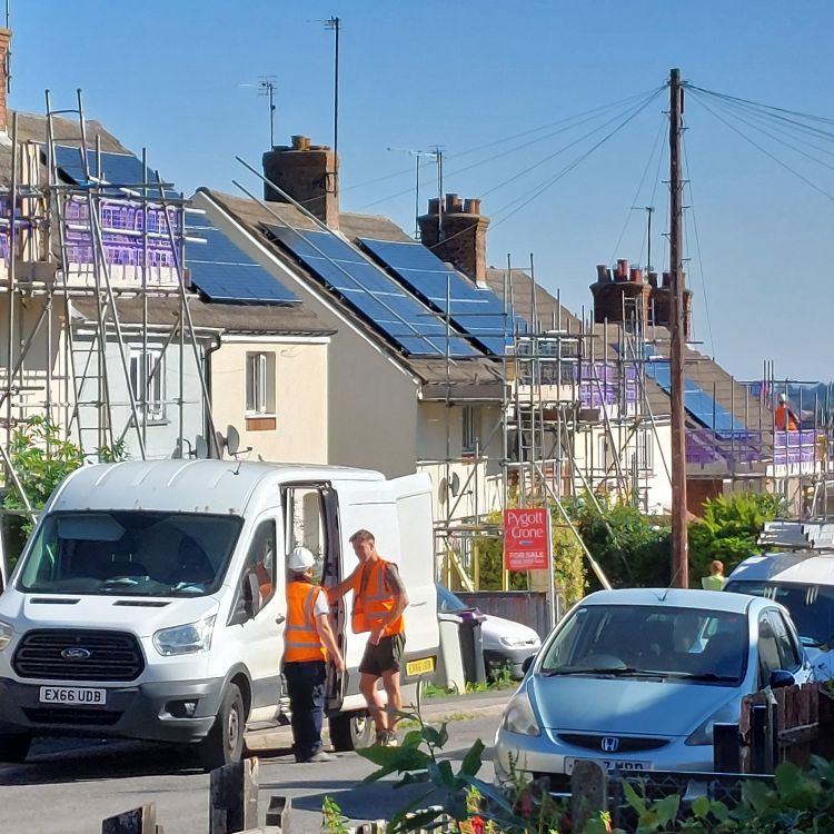 Construction workers beside a van on a residential street with scaffolding and solar panels being installed on rooftops.