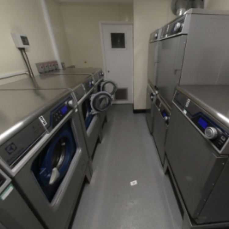 The laundry room at DeMontfort House with several stainless-steel commercial washing machines and dryers lined up on both sides, creating a central walkway towards a white door at the back.