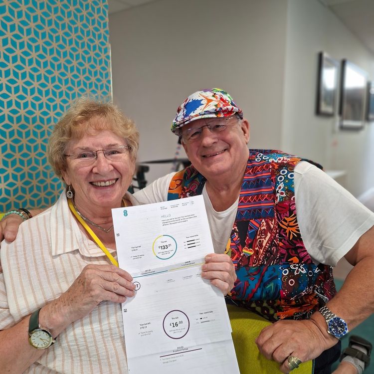 Annie, an older lady is sat down holding a bill showing a reduction in cost. Peter is kneeling next to her wearing a birght multi-coloured waistcoat and hat.