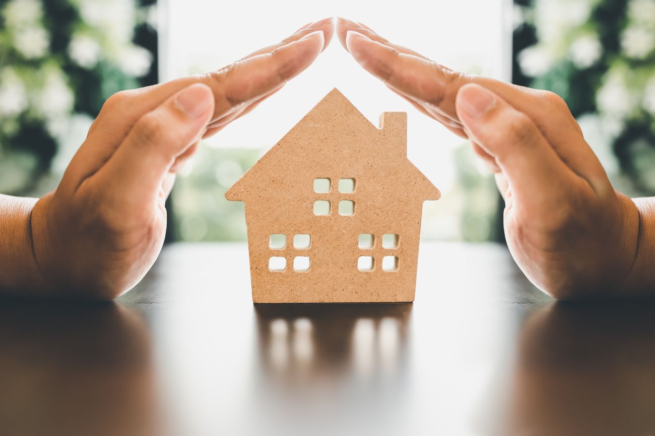 A cardboard cut-out of a house is held between two hands, with the fingers forming a protective roof shape above it. The background is softly blurred with natural green and white tones.