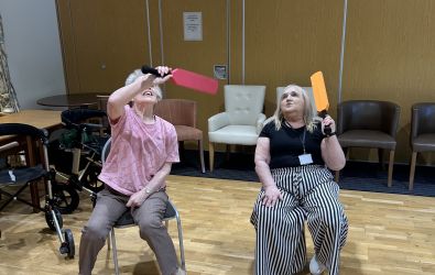 Two women seated in a community room play seated balloon cricket, holding foam cricket bats as a blue balloon floats above them. A mobility walker and chairs are visible in the background.
